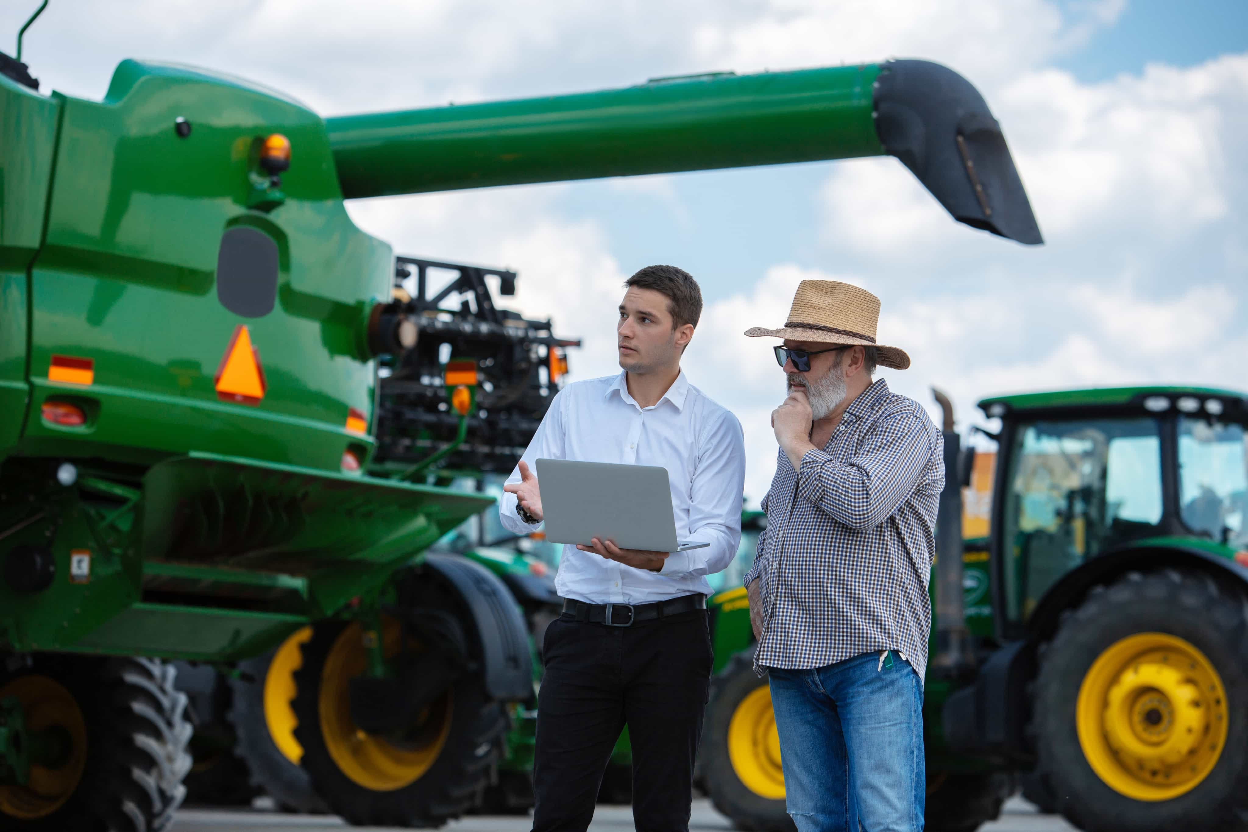 Business professional and farmer reviewing a laptop near agricultural equipment